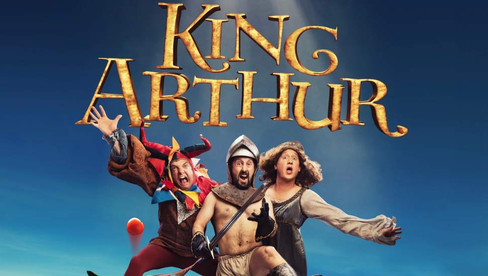 Promotional poster for a theatrical production of ‘King Arthur’ featuring three costumed characters in dynamic poses against a bright blue background. The title ‘King Arthur’ appears in large, ornate golden letters at the top
